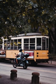 A bright yellow tram is moving along a tree-lined street, with lush green foliage overhead. In the foreground, a person rides a motorcycle, wearing a helmet and casual summer attire. The scene conveys urban movement, showcasing a mix of old and contemporary transportation.