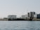 An industrial landscape with several large storage tanks and cylindrical structures near the shoreline. A rusty, partially sunken ship is located in the foreground, positioned along a rocky barrier. The sky is clear and the scene appears tranquil.