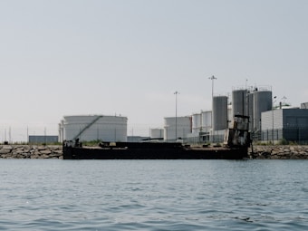 An industrial landscape with several large storage tanks and cylindrical structures near the shoreline. A rusty, partially sunken ship is located in the foreground, positioned along a rocky barrier. The sky is clear and the scene appears tranquil.