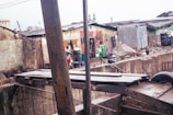 An urban slum area with makeshift houses, patched with corrugated metal sheets and wood. A child in a green shirt is walking through the area, carrying a bucket. Various household items, including buckets and pots, are scattered around. Clothes are hanging to dry on the walls of the buildings.