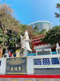 A public shrine featuring a large white statue of a deity, surrounded by vibrant mosaic tiles and traditional Chinese architectural elements. People are standing near the statue and trees line the background. A modern building is visible in the distance under a clear blue sky.