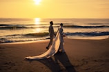 Wide shot of bride and groom walking hand in hand along a serene beach at sunset.