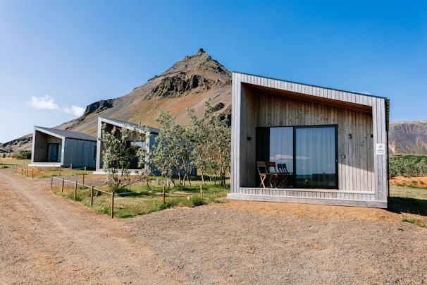 Several modern, minimalist cabins with wooden facades are positioned against a backdrop of a majestic, rocky mountain. The cabins have large glass windows and a simple, functional design. There is a dirt path leading to the cabins, and some trees and shrubs are scattered around the area. The sky is clear and blue, suggesting a sunny day.