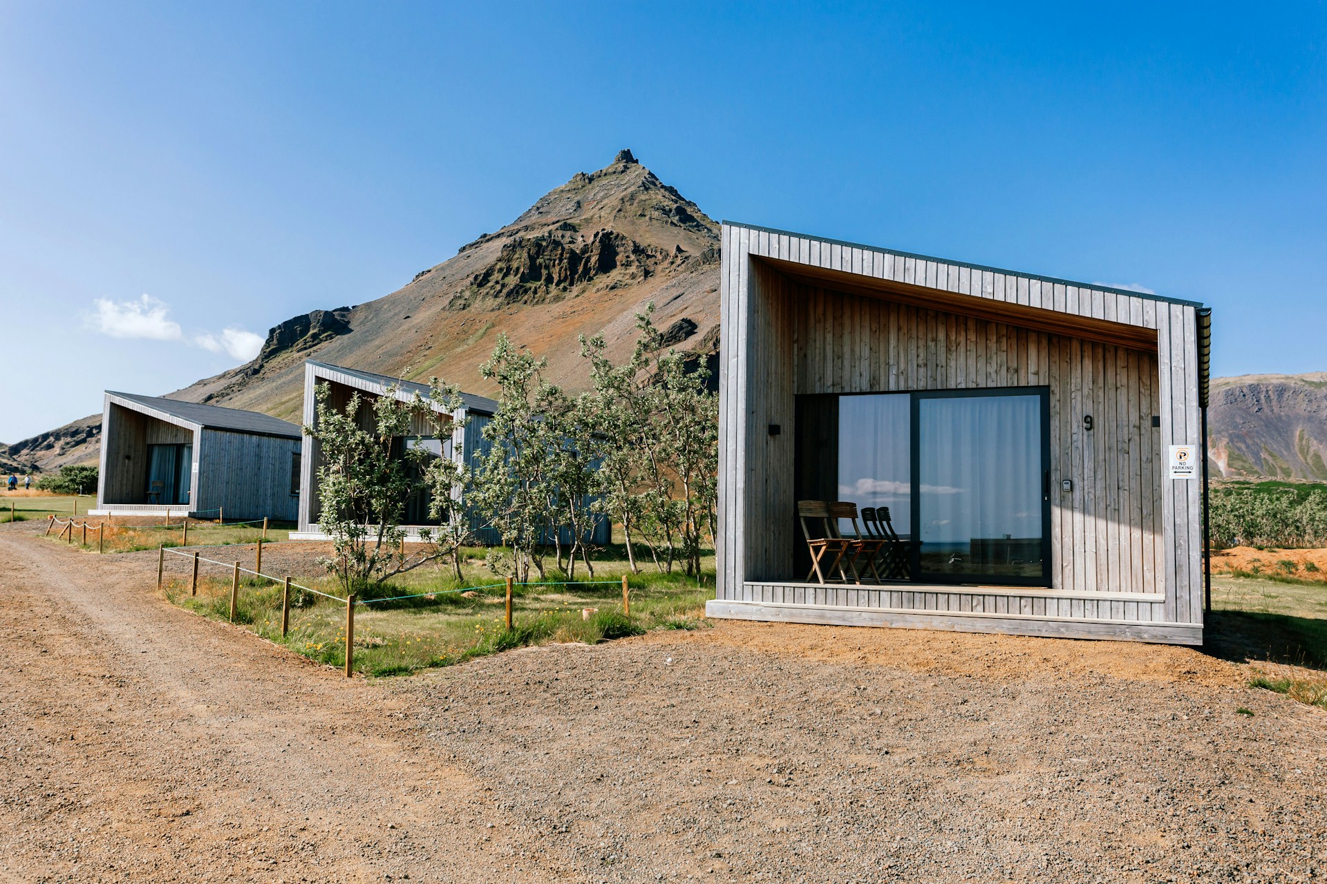 Several modern, minimalist cabins with wooden facades are positioned against a backdrop of a majestic, rocky mountain. The cabins have large glass windows and a simple, functional design. There is a dirt path leading to the cabins, and some trees and shrubs are scattered around the area. The sky is clear and blue, suggesting a sunny day.