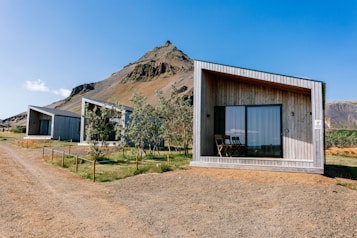Several modern, minimalist cabins with wooden facades are positioned against a backdrop of a majestic, rocky mountain. The cabins have large glass windows and a simple, functional design. There is a dirt path leading to the cabins, and some trees and shrubs are scattered around the area. The sky is clear and blue, suggesting a sunny day.