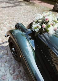 A vintage car with a glossy green finish is parked on a cobblestone path. Attached to the front of the car is a bouquet of pink and white flowers with lush greenery.