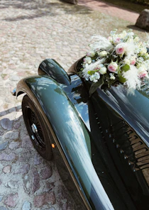 A vintage car with a glossy green finish is parked on a cobblestone path. Attached to the front of the car is a bouquet of pink and white flowers with lush greenery.