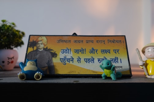 A decorative plaque with Hindi text featuring an image of a historical figure is centered on a desk. It is accompanied by small figurines, including two cartoon characters, one resembling a blue bear and the other a green turtle, as well as a small human figure in a yellow outfit holding a stick. In the background, there is a small potted plant in a decorative pot.