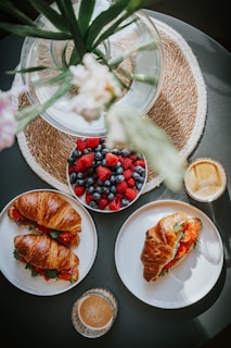A table set with a bowl of mixed berries including strawberries, blueberries, and raspberries. Two plates feature croissant sandwiches filled with greens and tomatoes. Two glasses of light brown coffee are also present. A decorative glass vase with green stems and a pink flower sits on a woven mat, creating a cozy and inviting brunch setting.