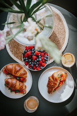 A table set with a bowl of mixed berries including strawberries, blueberries, and raspberries. Two plates feature croissant sandwiches filled with greens and tomatoes. Two glasses of light brown coffee are also present. A decorative glass vase with green stems and a pink flower sits on a woven mat, creating a cozy and inviting brunch setting.