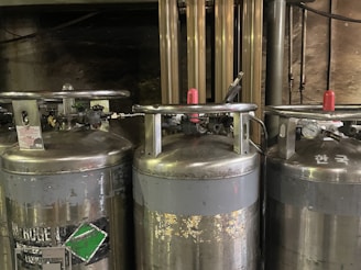 Three large industrial gas cylinders with metallic surfaces are aligned side by side. They have pressure gauges, valves, and connecting hoses attached to the top. The cylinders have some wear and tear visible, with labels and signs of use. The background shows pipes and a dark, industrial setting.