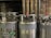 Three large industrial gas cylinders with metallic surfaces are aligned side by side. They have pressure gauges, valves, and connecting hoses attached to the top. The cylinders have some wear and tear visible, with labels and signs of use. The background shows pipes and a dark, industrial setting.