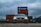 A grey Volkswagen car is prominently positioned in the foreground with its headlights on. Behind the car, there are stacked shipping containers in various colors, prominently featuring branding. The sky is overcast, creating a moody atmosphere.