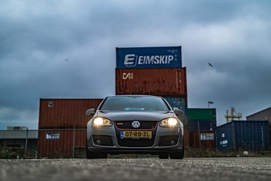 A grey Volkswagen car is prominently positioned in the foreground with its headlights on. Behind the car, there are stacked shipping containers in various colors, prominently featuring branding. The sky is overcast, creating a moody atmosphere.
