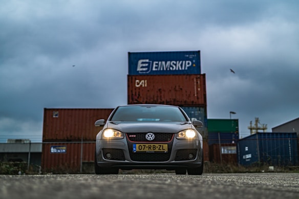 A grey Volkswagen car is prominently positioned in the foreground with its headlights on. Behind the car, there are stacked shipping containers in various colors, prominently featuring branding. The sky is overcast, creating a moody atmosphere.