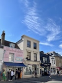 A street view featuring a series of charming storefronts under a bright blue sky. The first building displays a pastel pink exterior with the name 'Wheelers Oyster Bar' and an illustration of seafood. People are gathered by the entrance. Next door, a larger white building houses a shop labeled 'djl Hair & Beauty'. The street is lively with pedestrians strolling along the sidewalk.