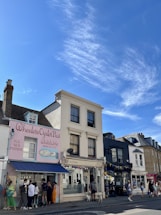 A street view featuring a series of charming storefronts under a bright blue sky. The first building displays a pastel pink exterior with the name 'Wheelers Oyster Bar' and an illustration of seafood. People are gathered by the entrance. Next door, a larger white building houses a shop labeled 'djl Hair & Beauty'. The street is lively with pedestrians strolling along the sidewalk.