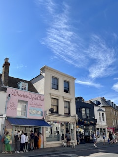 A street view featuring a series of charming storefronts under a bright blue sky. The first building displays a pastel pink exterior with the name 'Wheelers Oyster Bar' and an illustration of seafood. People are gathered by the entrance. Next door, a larger white building houses a shop labeled 'djl Hair & Beauty'. The street is lively with pedestrians strolling along the sidewalk.