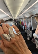 A close-up of a hand wearing two rings, one shaped like a butterfly, inside an airplane cabin. The background is slightly blurred showing rows of occupied seats and overhead luggage compartments.