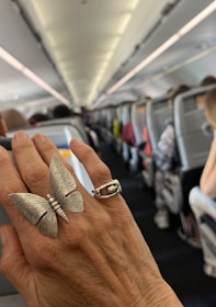 A close-up of a hand wearing two rings, one shaped like a butterfly, inside an airplane cabin. The background is slightly blurred showing rows of occupied seats and overhead luggage compartments.