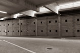 A large, empty parking garage with a concrete ceiling and walls featuring small square windows. The floor is paved with cobblestones, and there is ambient lighting coming from the ceiling fixtures.