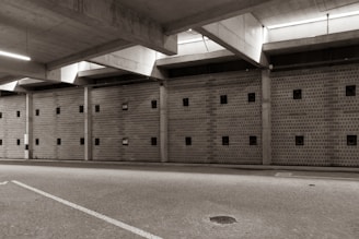 A large, empty parking garage with a concrete ceiling and walls featuring small square windows. The floor is paved with cobblestones, and there is ambient lighting coming from the ceiling fixtures.
