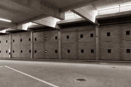 A large, empty parking garage with a concrete ceiling and walls featuring small square windows. The floor is paved with cobblestones, and there is ambient lighting coming from the ceiling fixtures.