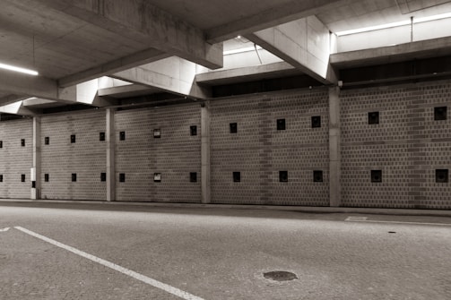 A large, empty parking garage with a concrete ceiling and walls featuring small square windows. The floor is paved with cobblestones, and there is ambient lighting coming from the ceiling fixtures.