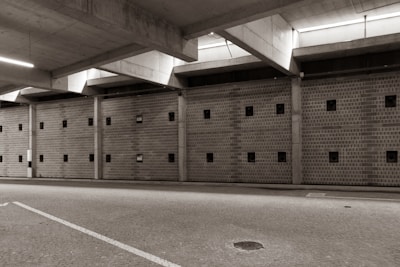 A large, empty parking garage with a concrete ceiling and walls featuring small square windows. The floor is paved with cobblestones, and there is ambient lighting coming from the ceiling fixtures.