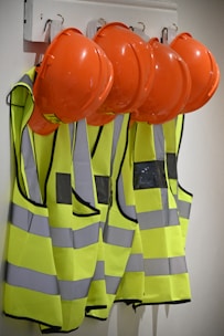 Several bright orange hard hats and fluorescent yellow safety vests with reflective strips are hanging on hooks on a wall.