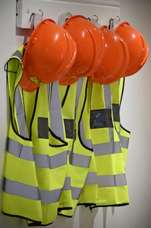 Several bright orange hard hats and fluorescent yellow safety vests with reflective strips are hanging on hooks on a wall.