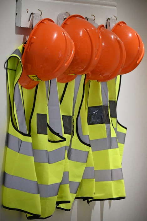 Several bright orange hard hats and fluorescent yellow safety vests with reflective strips are hanging on hooks on a wall.
