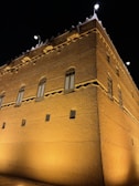 A traditional mud-brick building is captured at night, with intricate patterns on its walls and several shuttered windows. The structure is illuminated by bright yellow lights, highlighting its textured surface. The rooftop features triangular spikes, and there are modern street lamps on both corners of the building.