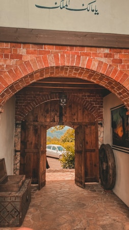 An arched brick entrance leads to a wooden door that is ajar, revealing a view of a parked white car and lush greenery outside. The stone floor and walls enhance the rustic ambiance, and there is an old wooden chest and a large wooden wheel inside the entrance. Above the door, an Arabic inscription decorates the wall.