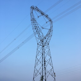 A large electricity transmission tower with a unique twisted design extends into the sky. Multiple power lines are connected along the tower's frame, stretching horizontally across the clear, blue sky.