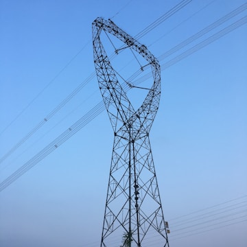 A large electricity transmission tower with a unique twisted design extends into the sky. Multiple power lines are connected along the tower's frame, stretching horizontally across the clear, blue sky.