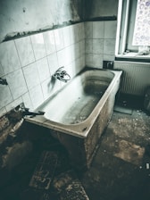 An old, worn-out bathtub is set against a wall with white tiles. The bathroom appears neglected, with visible signs of decay and grime. A single window partially covered with a decorative film allows some light to enter, revealing the aged state of the room.