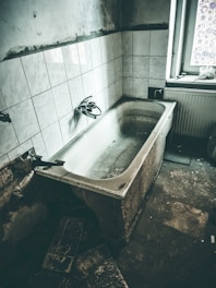 An old, worn-out bathtub is set against a wall with white tiles. The bathroom appears neglected, with visible signs of decay and grime. A single window partially covered with a decorative film allows some light to enter, revealing the aged state of the room.