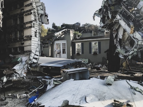 Debris from a destroyed structure is scattered around a partially collapsed building. Wreckage, including torn metal and broken panels, surrounds the scene. The building itself, with exposed interiors, stands amid the chaos of scattered materials.