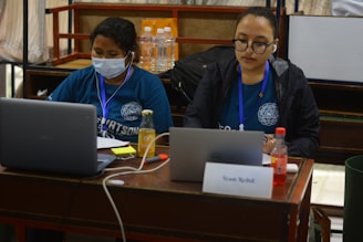 Two individuals are sitting at a table working on laptops. One person is wearing a mask and a blue shirt, while the other is wearing glasses and a black jacket, both with lanyards. There are bottles of water and juice on the table along with a card labeled 'Team Rebuilt'.