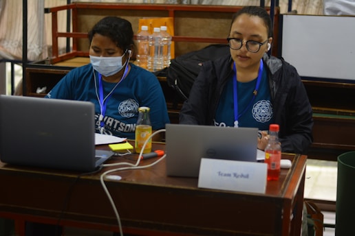 Two individuals are sitting at a table working on laptops. One person is wearing a mask and a blue shirt, while the other is wearing glasses and a black jacket, both with lanyards. There are bottles of water and juice on the table along with a card labeled 'Team Rebuilt'.