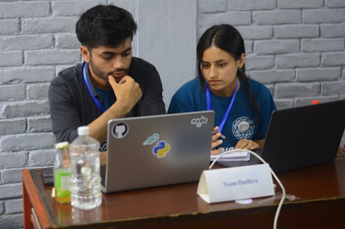 Two people appear focused and engaged while working on laptops at a wooden table. Both are wearing badge lanyards and seem to be collaborating. The table includes bottled beverages, a notebook, and a name plate that reads 'Team Outliers'. Stickers on one laptop suggest topics related to programming or software development.