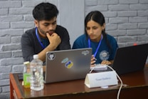 Two people appear focused and engaged while working on laptops at a wooden table. Both are wearing badge lanyards and seem to be collaborating. The table includes bottled beverages, a notebook, and a name plate that reads 'Team Outliers'. Stickers on one laptop suggest topics related to programming or software development.