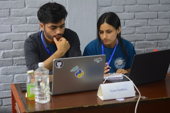 Two people appear focused and engaged while working on laptops at a wooden table. Both are wearing badge lanyards and seem to be collaborating. The table includes bottled beverages, a notebook, and a name plate that reads 'Team Outliers'. Stickers on one laptop suggest topics related to programming or software development.
