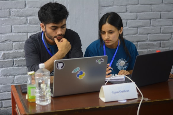 Two people appear focused and engaged while working on laptops at a wooden table. Both are wearing badge lanyards and seem to be collaborating. The table includes bottled beverages, a notebook, and a name plate that reads 'Team Outliers'. Stickers on one laptop suggest topics related to programming or software development.