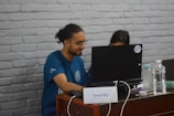 A person with long hair tied back is sitting at a desk and looking at a laptop. Next to the laptop, there is a plastic water bottle and another beverage container. The person is wearing a blue t-shirt and is accompanied by another individual partially visible behind the laptop. The desk has a small sign labeled 'Team ATGC.' The background features a white brick wall.