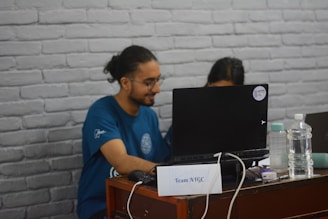 A person with long hair tied back is sitting at a desk and looking at a laptop. Next to the laptop, there is a plastic water bottle and another beverage container. The person is wearing a blue t-shirt and is accompanied by another individual partially visible behind the laptop. The desk has a small sign labeled 'Team ATGC.' The background features a white brick wall.
