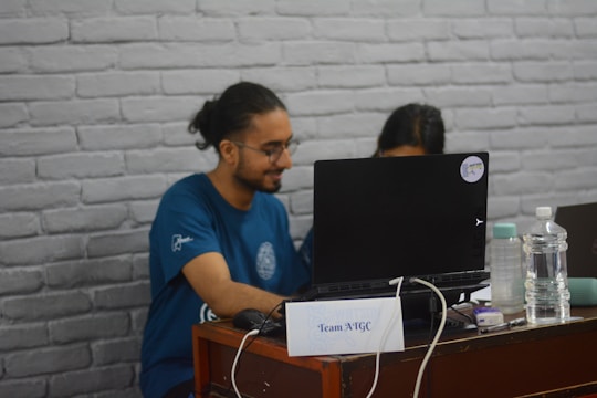 A person with long hair tied back is sitting at a desk and looking at a laptop. Next to the laptop, there is a plastic water bottle and another beverage container. The person is wearing a blue t-shirt and is accompanied by another individual partially visible behind the laptop. The desk has a small sign labeled 'Team ATGC.' The background features a white brick wall.