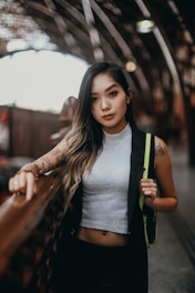 A young woman with tattoos stands beside an ornate railing in a dimly lit setting. She is wearing a sleeveless top and has long hair with a gradient of colors. The background shows an out-of-focus architectural structure with curved elements and bright lights.