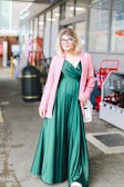A person in a flowing emerald green dress, complemented by a stylish pink blazer. The individual is standing in an urban environment, holding a small white handbag. The background includes elements such as a trash bin, shopping carts, and a storefront.
